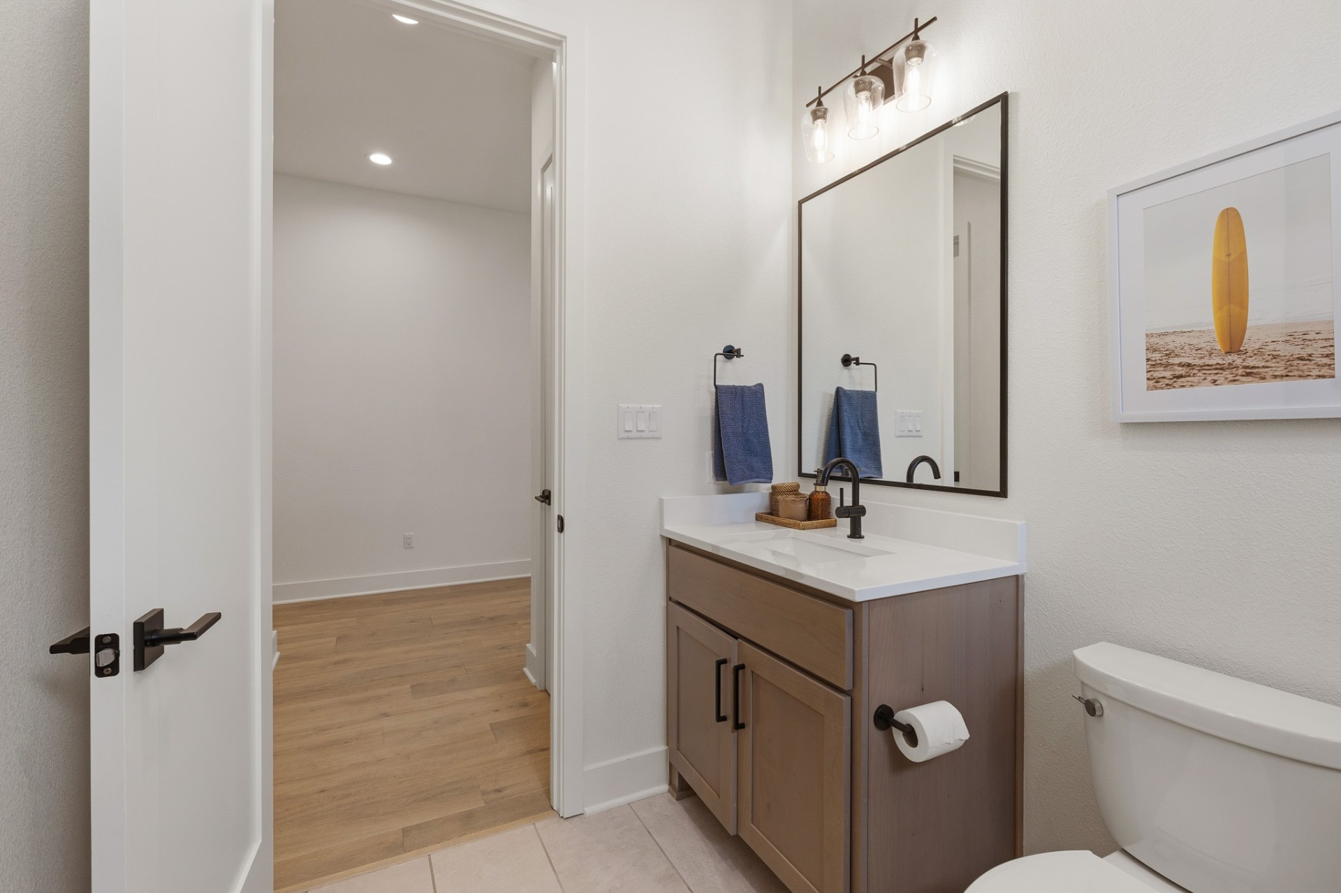 Half bath vanity — tan shaker cabinet, black-frame mirror, surfboard art and hallway view
